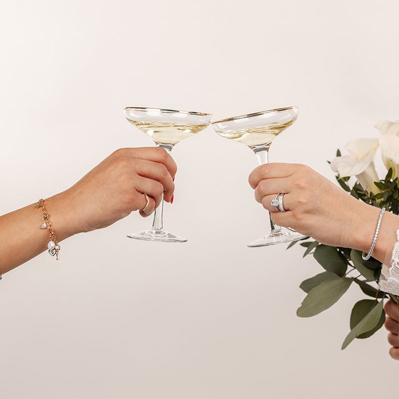 Zwei Frauen tragen funkelnde Ringe und Armbänder von Amor und stoßen auf einer Hochzeit miteinander an
